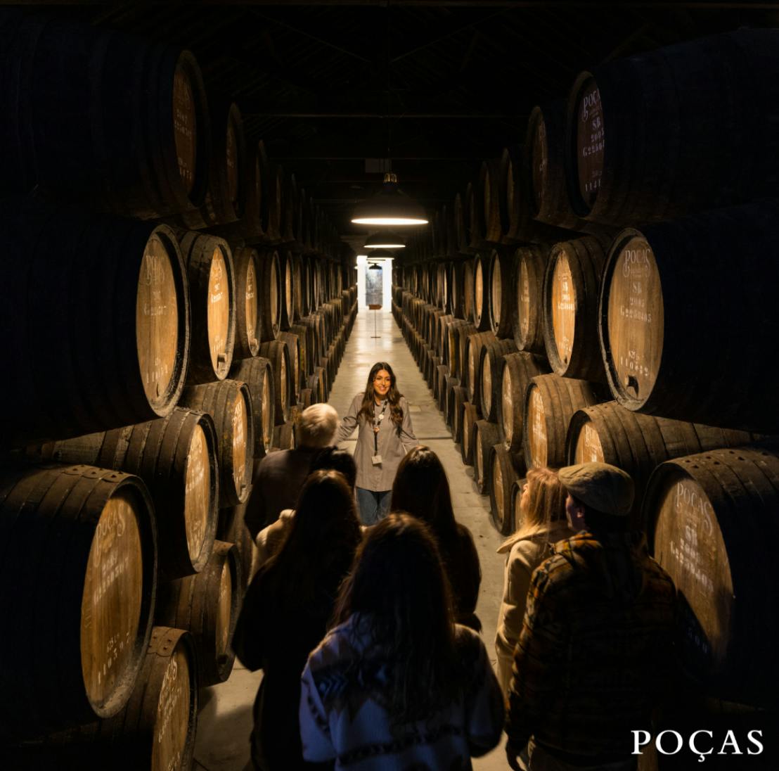 Un grupo de personas en una visita guiada al interior de una bodega con hileras de barriles de madera a ambos lados.