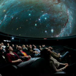 Människor tittar på en rymdsimulering i ett mörkt planetarium, sitter och tittar upp på en projicerad galax på ett kupoltak.
