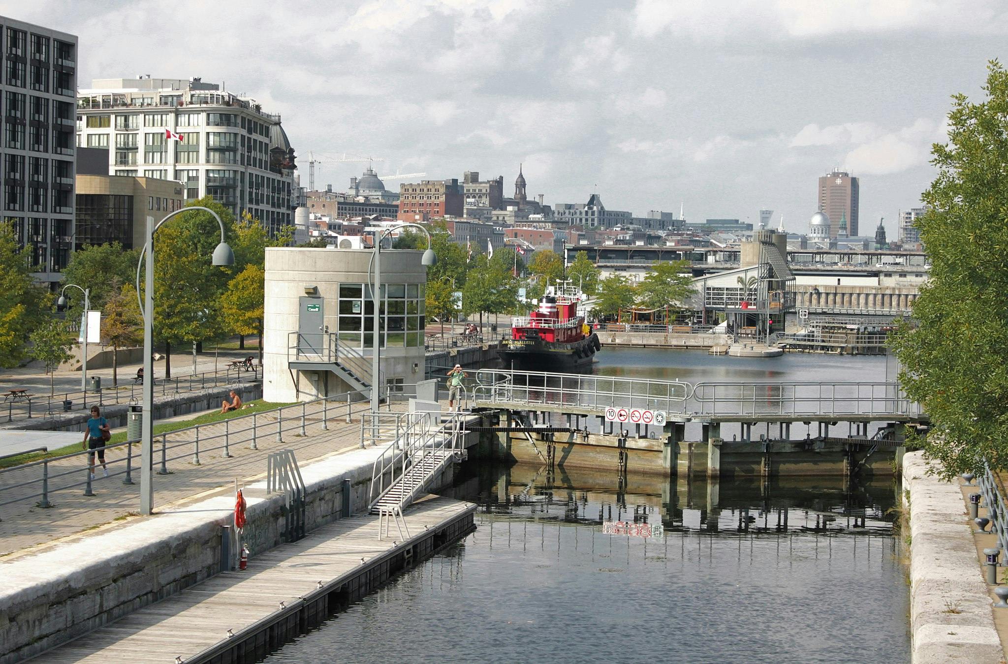 Urban riverside scene with red boat, walkway, small building, and city skyline backdrop. People strolling and relaxing nearby.