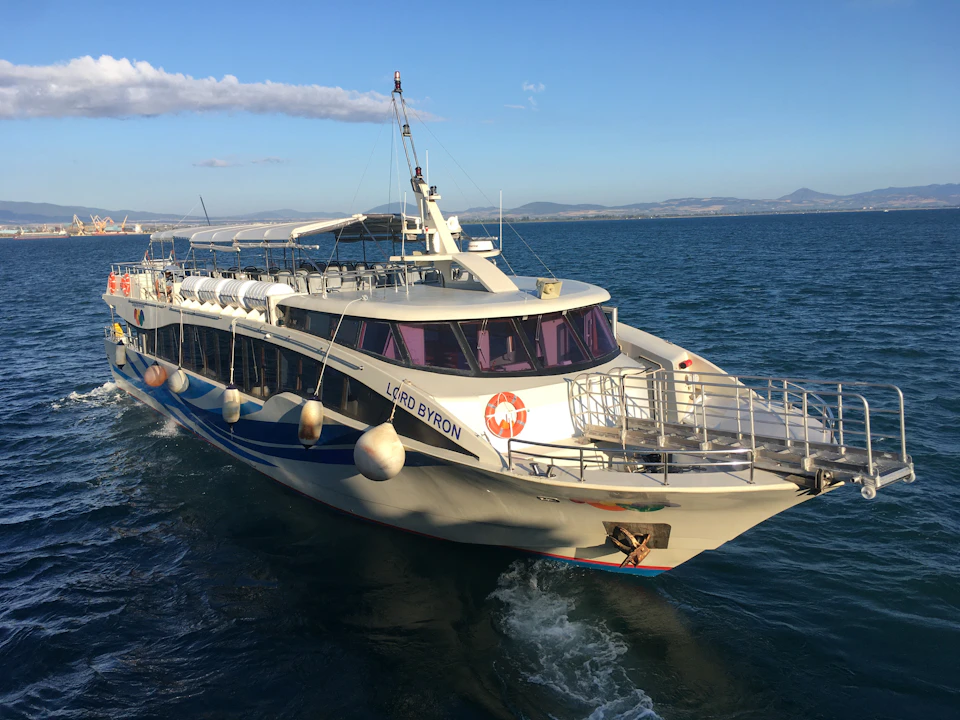 A white passenger ferry named 'Lord Byron' navigates through calm blue waters under a clear sky. Mountains are visible in the background.