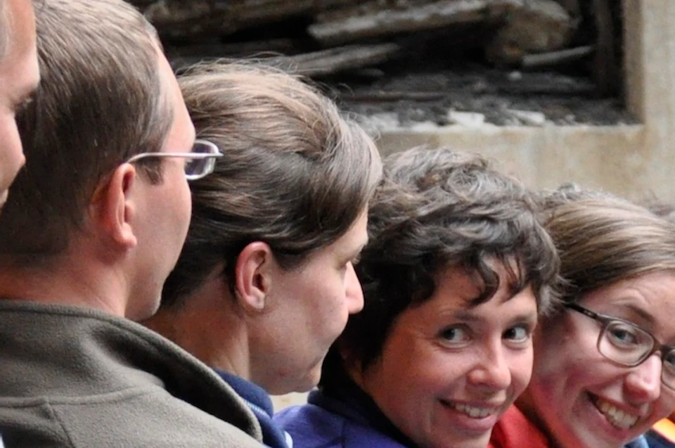 A group of people stand in line indoors, facing a screen displaying their own image. Rubble and damaged walls are visible in the background.