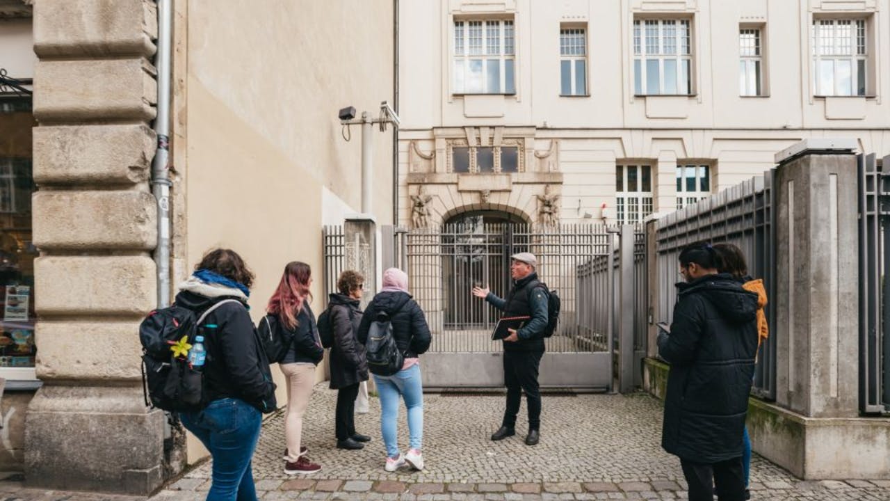 A tour guide gesturing while four people with backpacks listen in a cobblestone alley next to a historic building.