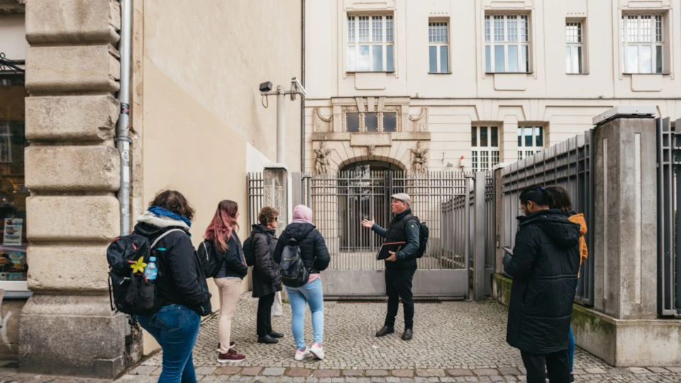 A tour guide gesturing while four people with backpacks listen in a cobblestone alley next to a historic building.