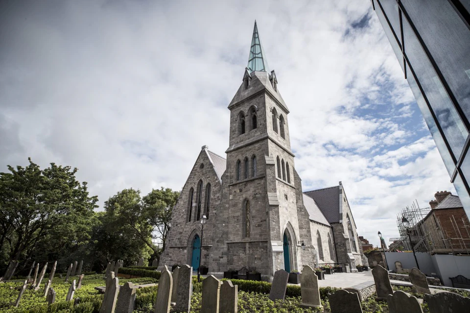 Stone church with a tall spire, blue doors, and surrounding gravestones under a partly cloudy sky. Trees are visible in the background.