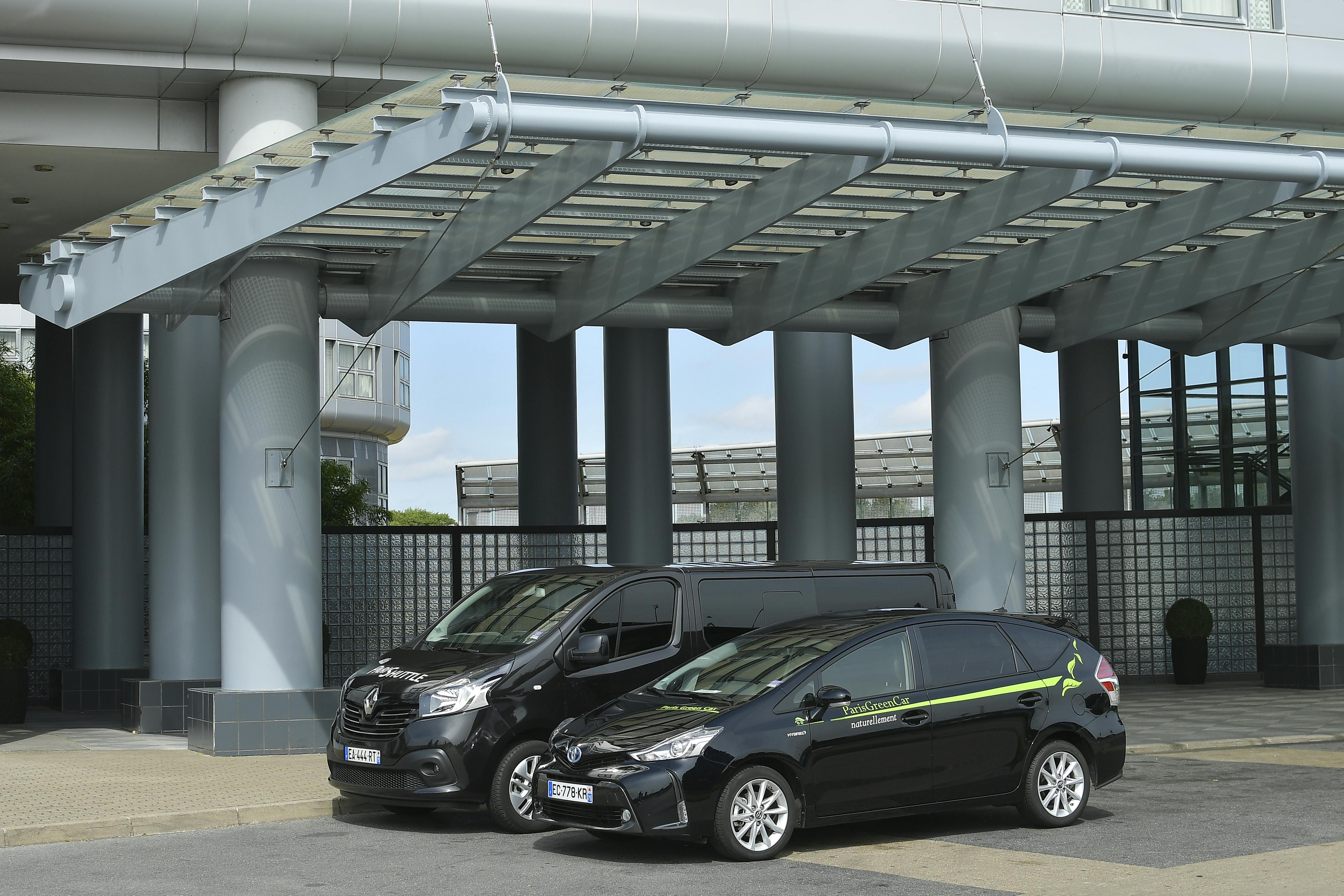 スチールキャノピーの下に駐車された2台の黒い車両、1台はバン、もう1台は緑色の「タクシーグリーン」テキストのセダン。
