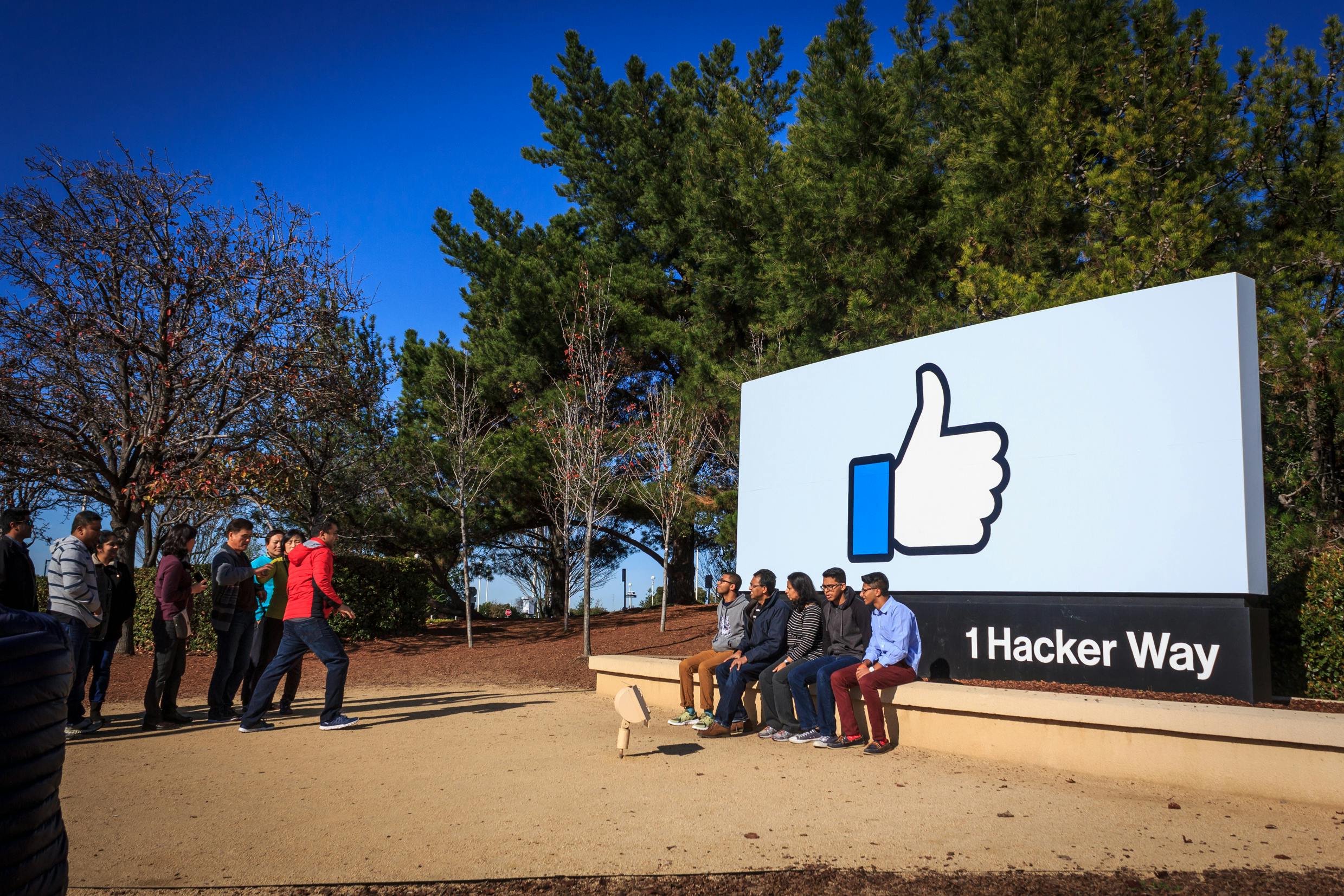 Persone che posano e scattano foto vicino a un grande cartello con l'icona del pollice in su e il testo "1 Hacker Way" circondato da alberi.