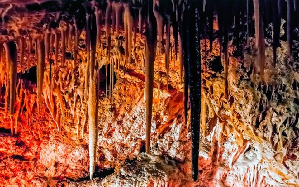 Stalactites and stalagmites in a dimly lit cave with shades of red and orange on the rock formations.