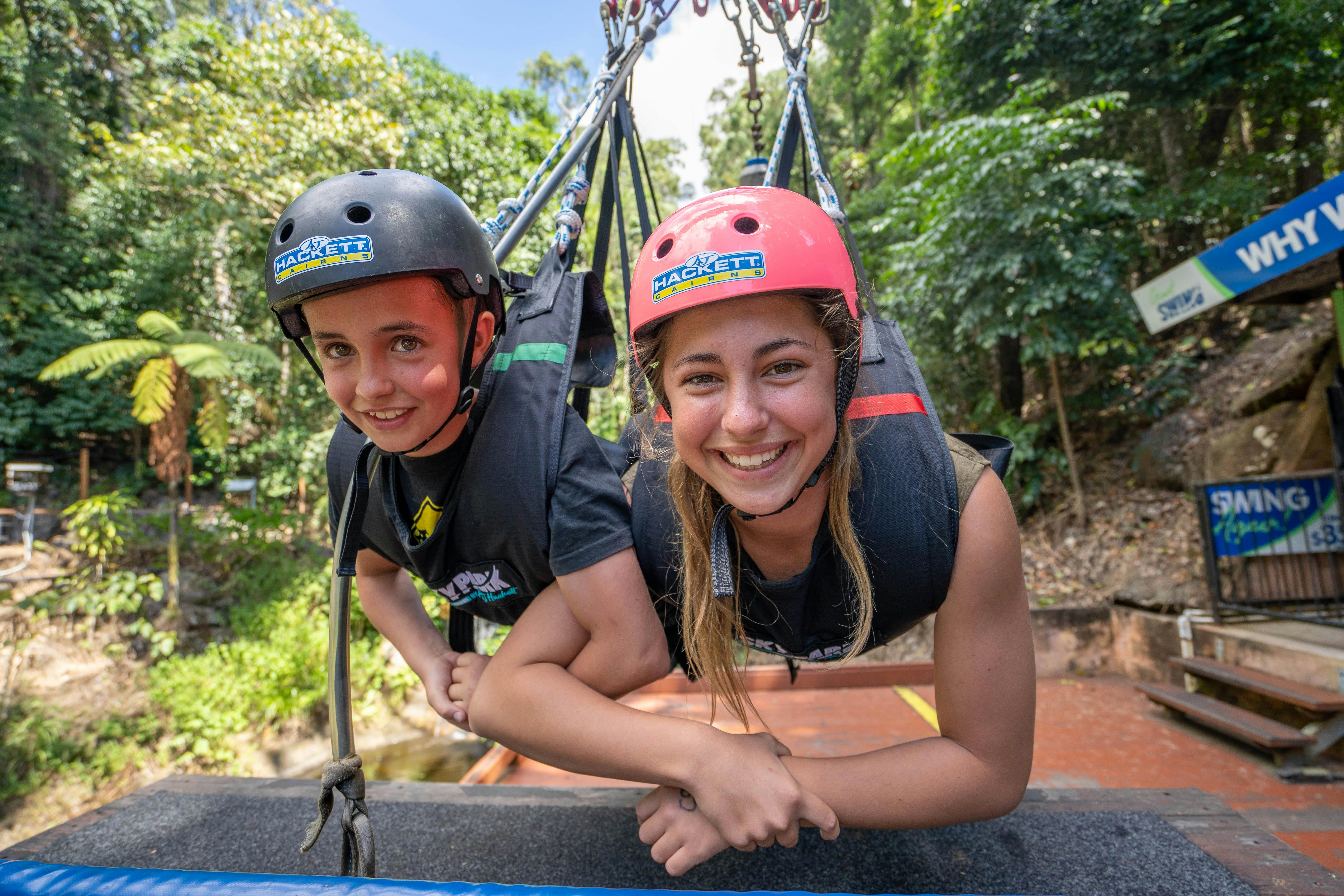 两个戴着头盔的人，微笑着牵着手，悬挂在郁郁葱葱的森林户外环境中。