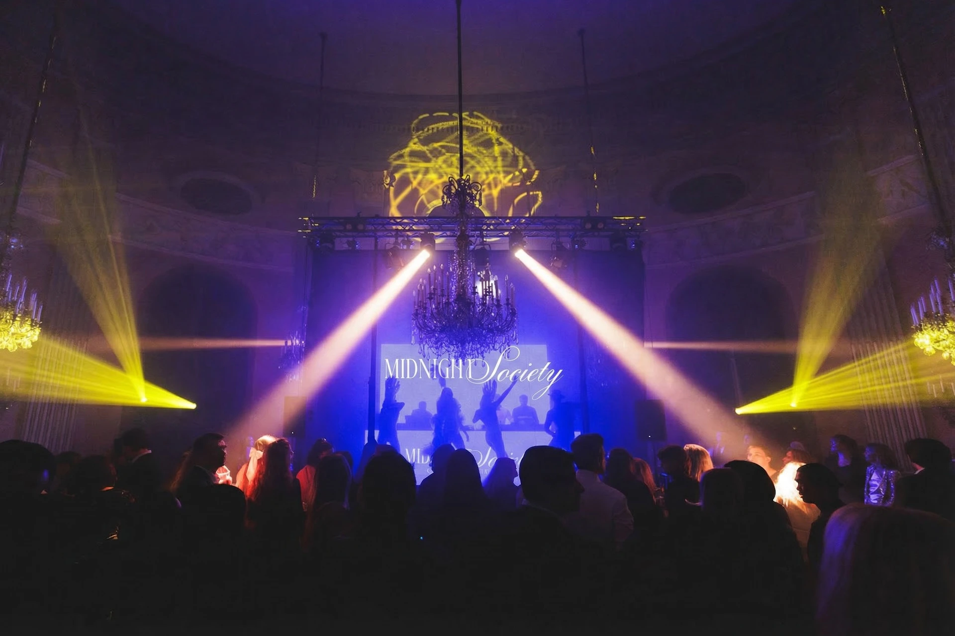 A dimly lit event with people gathered, stage lights, a chandelier, and a screen displaying 'Midnight Society.'