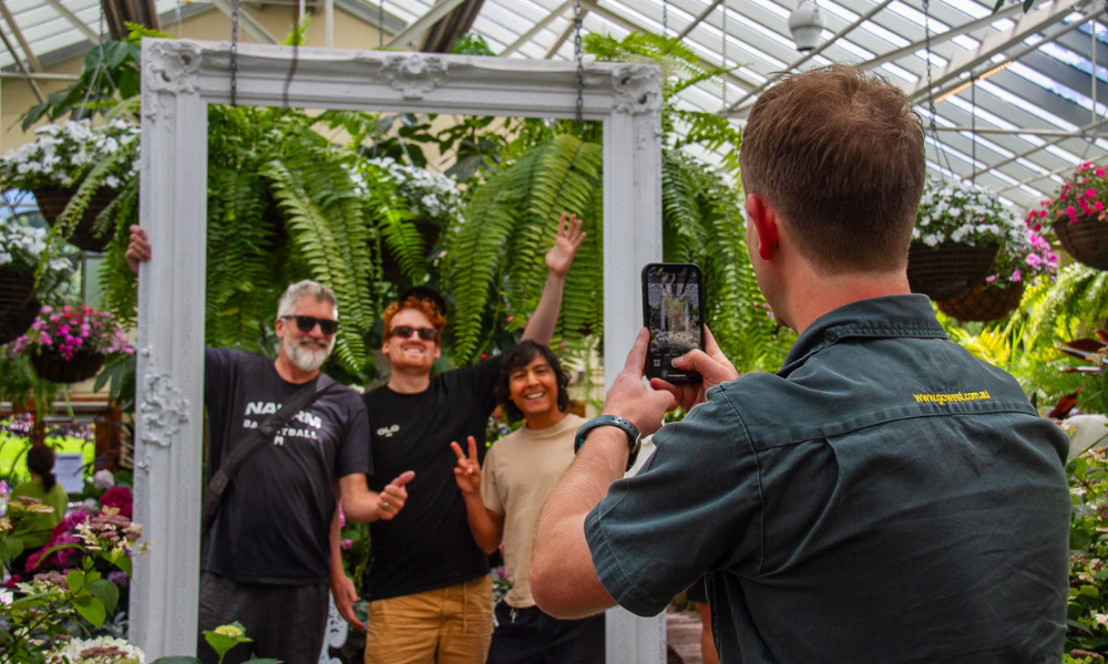 Three men posing inside a large, white frame surrounded by greenery, while another person photographs them with a smartphone.