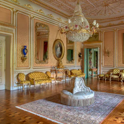 Ett utsmyckat, antikt rum med en stor ljuskrona, guldklädda möbler, porträtt och en detaljerad matta på ett parkettgolv.