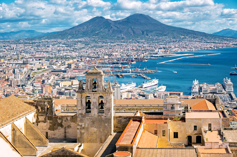 A cityscape with a historic bell tower in the foreground, a harbor with ships, and a mountain under a blue sky in the distance.
