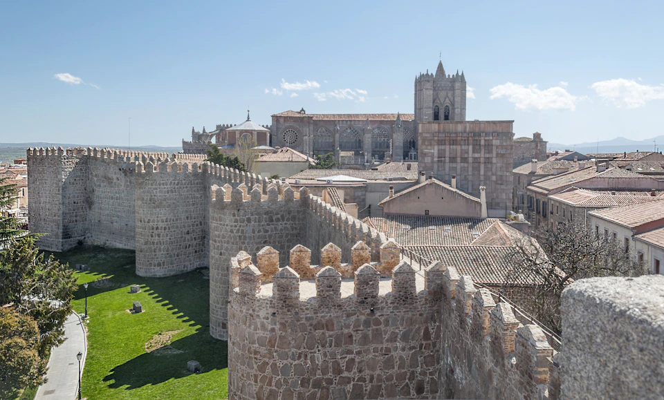 Stone city walls with watchtowers surround medieval buildings and a large cathedral under a clear blue sky.