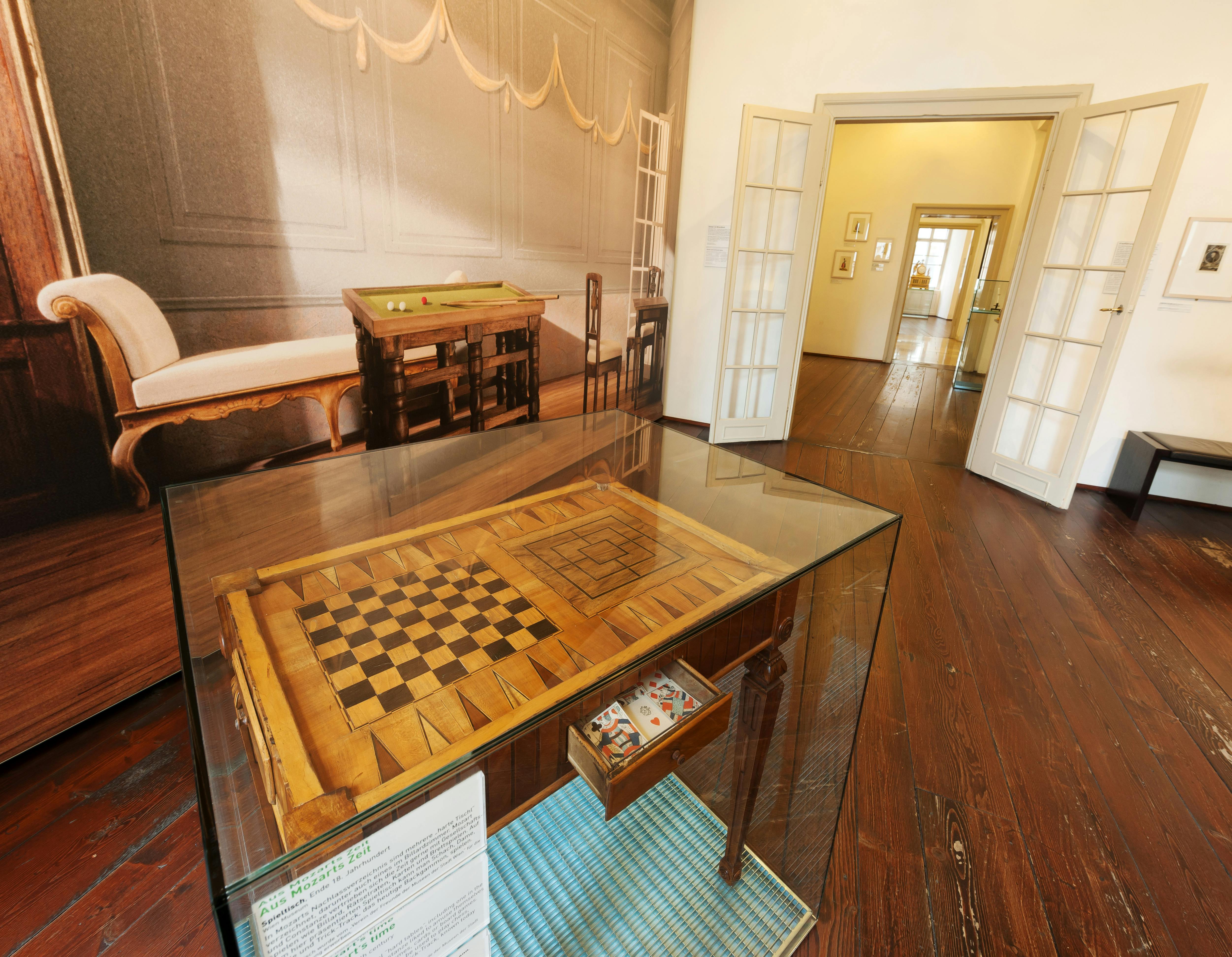 In een kamer met houten vloeren staat een schaaktafel in een glazen kast. Een aangrenzende kamer is zichtbaar door een open deuropening.