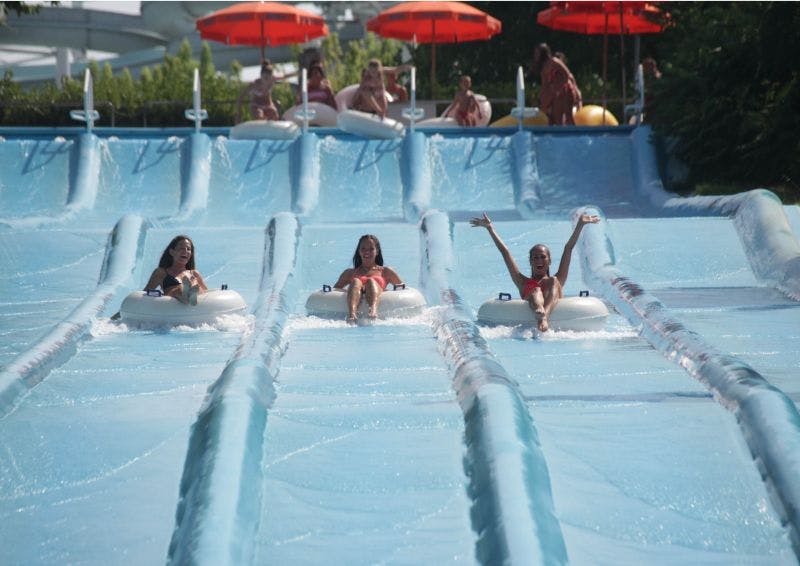 Three women in swimsuits ride down a water slide in inflatable tubes, with several others waiting at the top under red umbrellas.