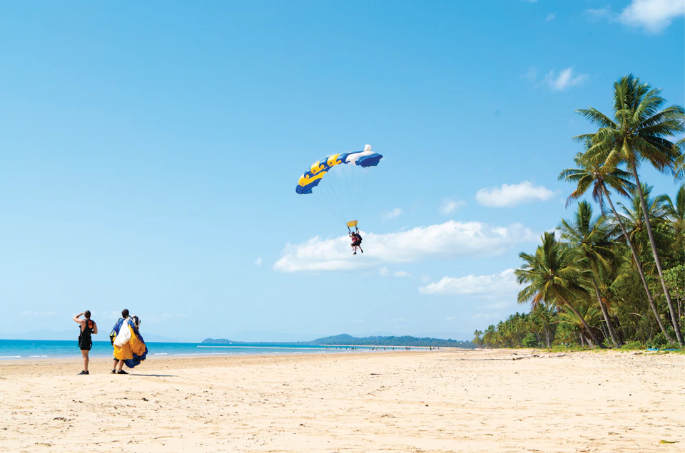A person parasailing over a sandy beach, with a yellow and blue parachute. Another person in the foreground looks on.