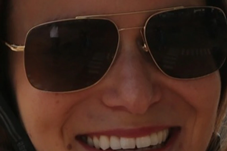 A smiling person wearing a red helmet and sunglasses outdoors with a blurred architectural structure in the background.