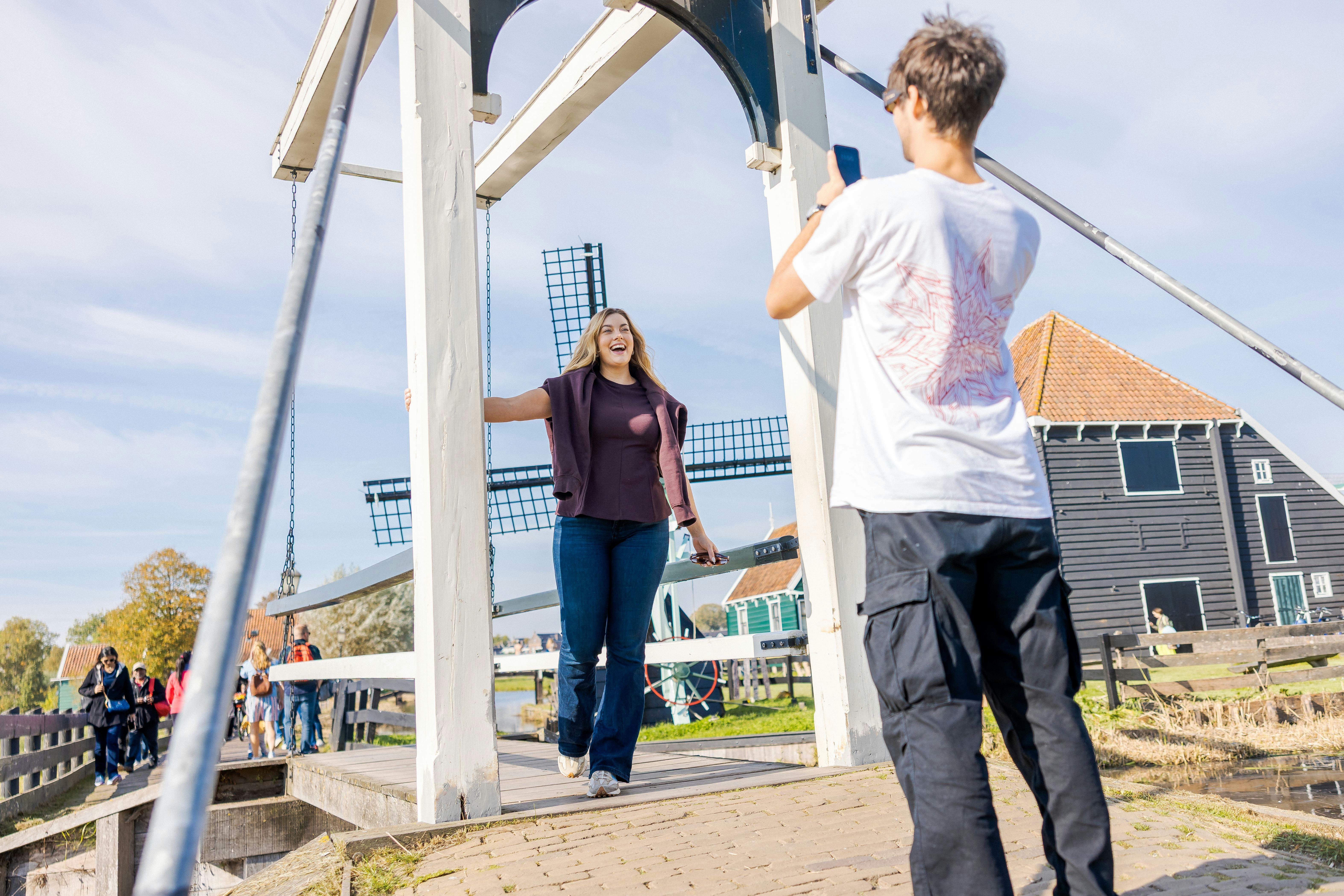 A person photographs a smiling woman posing under a white bridge with a windmill and buildings in the background.