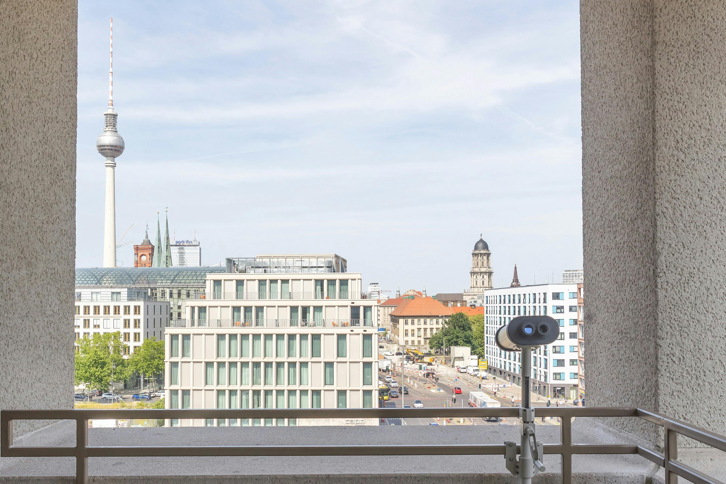 A cityscape view from a balcony showing modern and historic buildings, with a binocular viewer in the foreground.