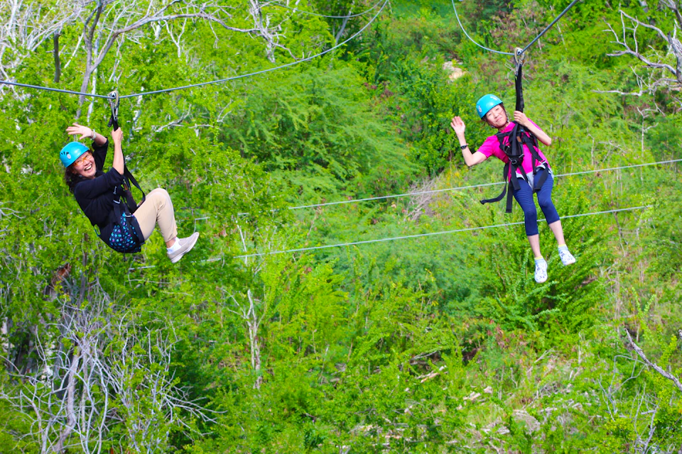 Two people ziplining over a lush green forest, one waving while wearing a helmet and safety gear.