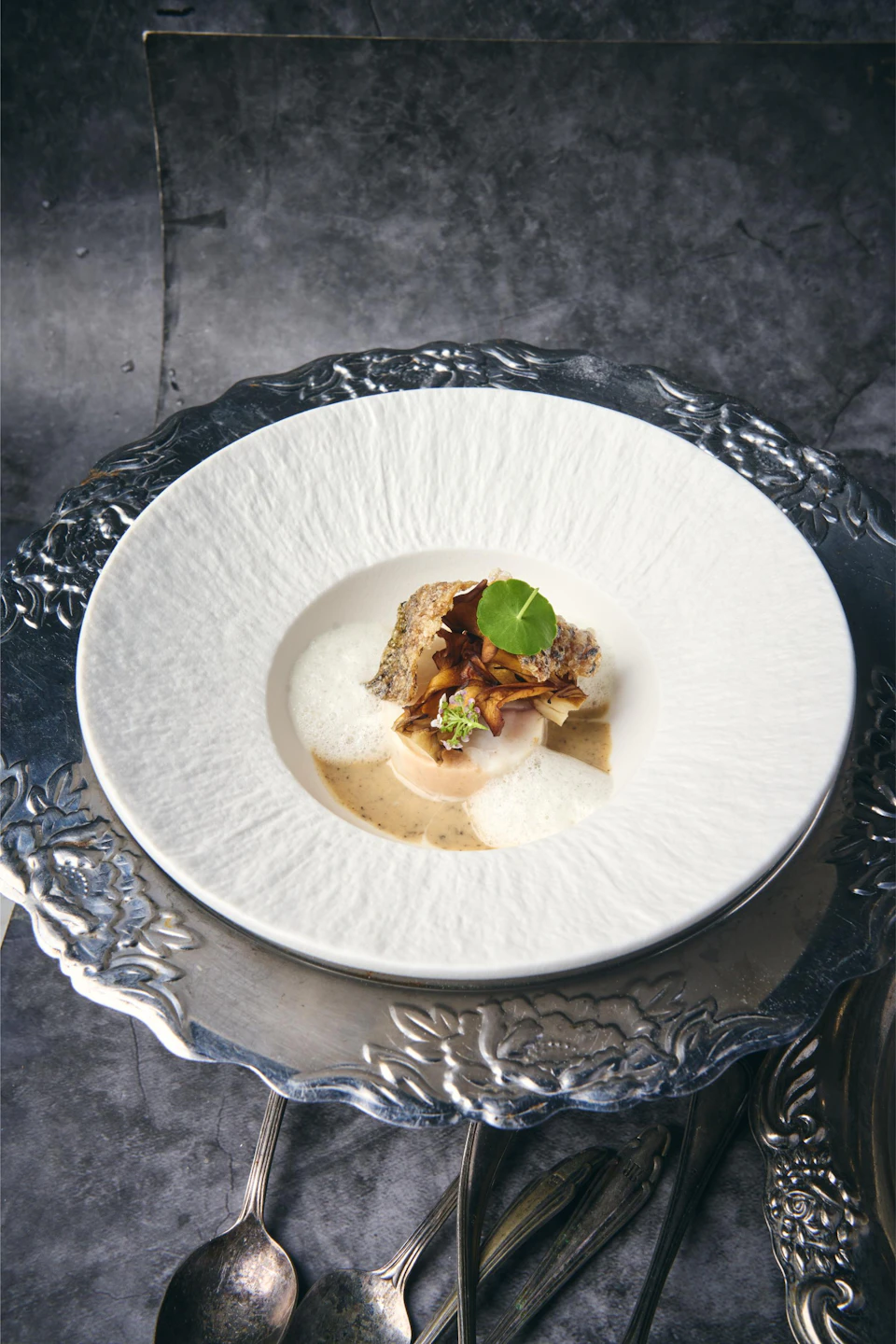 A decorative plate with a gourmet dish, including foam, herbs, and a crispy garnish, presented on an ornate silver tray.