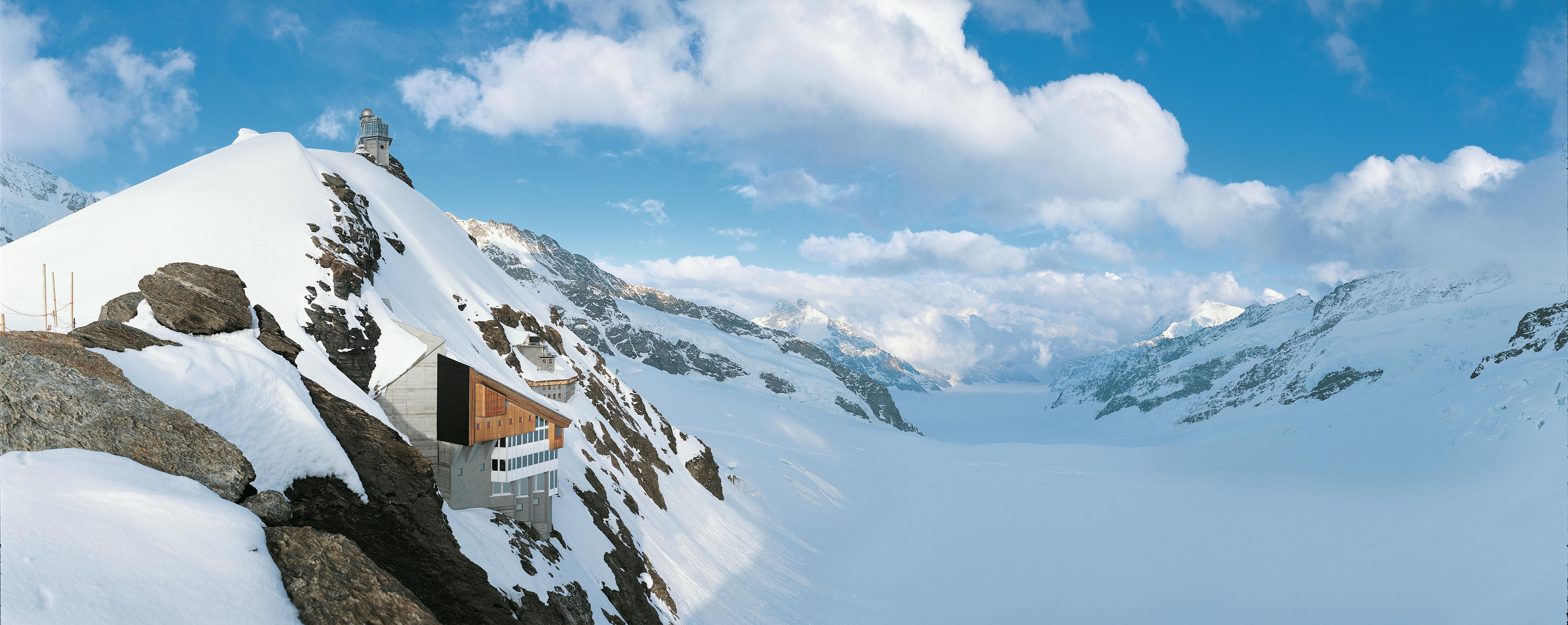 部分的に曇った空の下、斜面に木造の建物がある雪に覆われた山々。