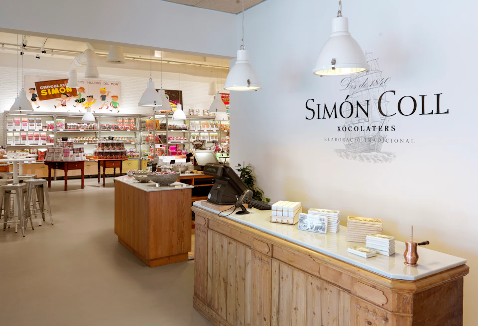 A shop interior with a wooden counter, cash register, hanging lights, shelves of chocolate products, and a sign reading "Simón Con."