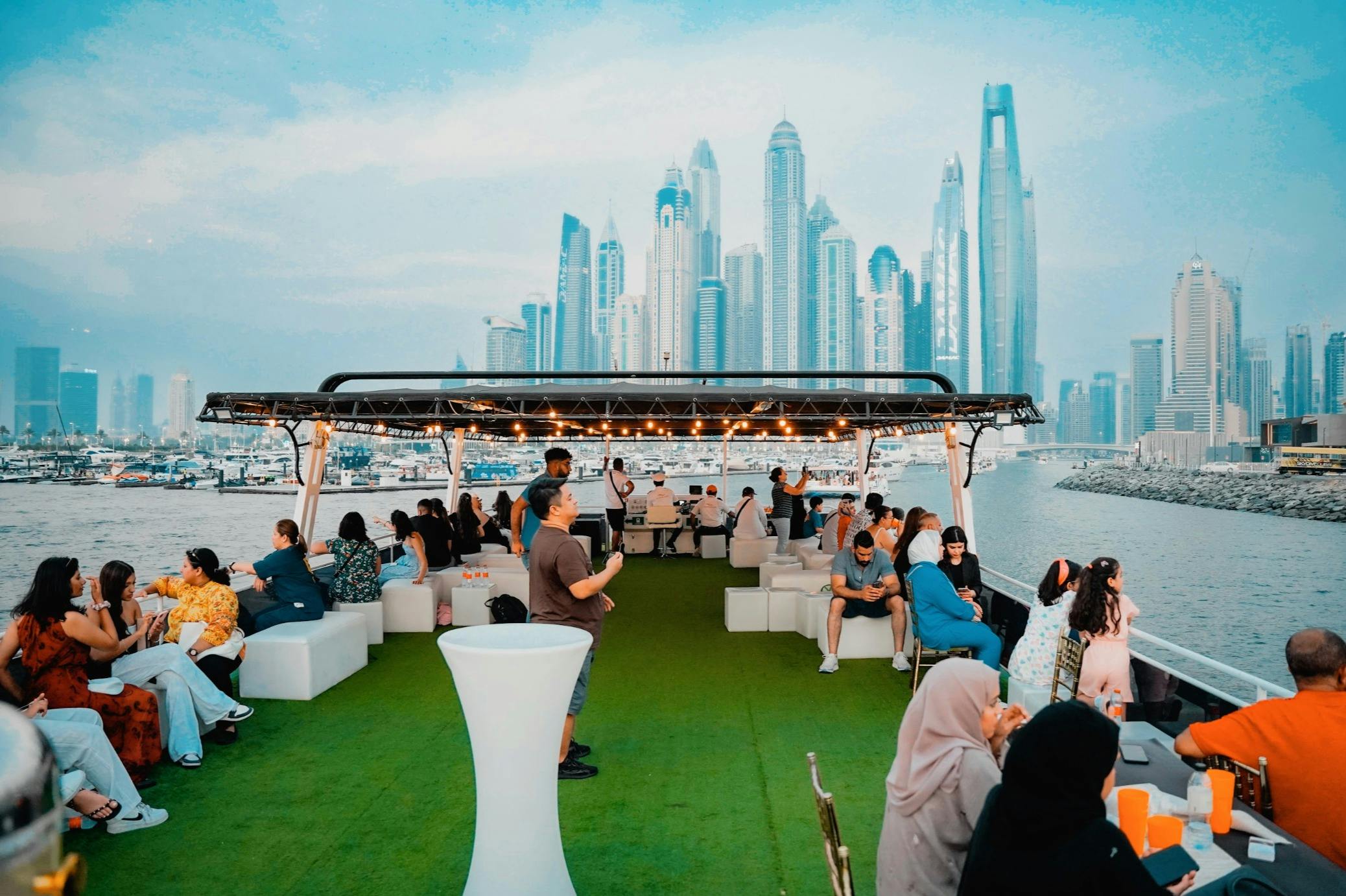 Människor sitter på vita soffor och pallar på en båt med konstgräsgolv, med en stadsbild av höga byggnader i bakgrunden.