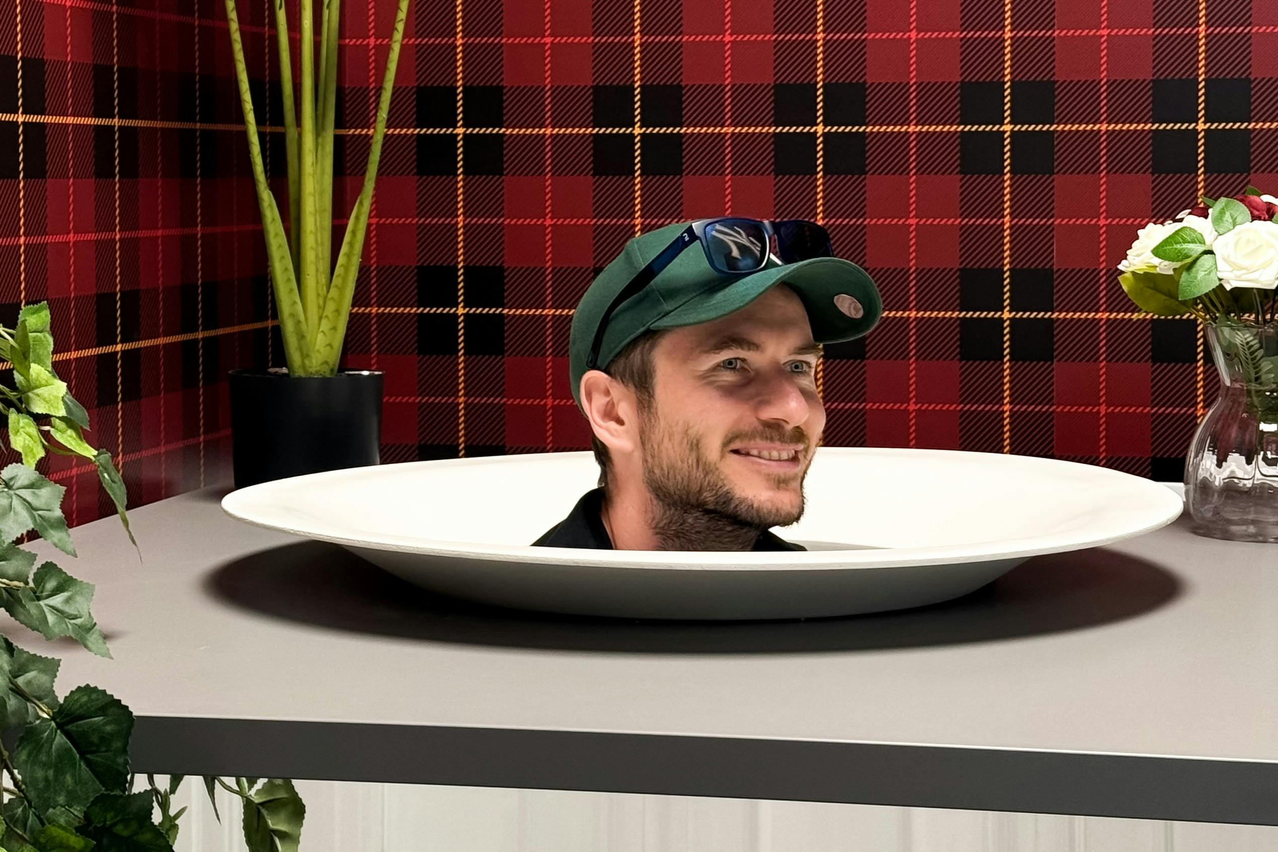 En mans huvud visas på en tallrik på ett bord, omgivet av växter och en röd rutig bakgrund.
