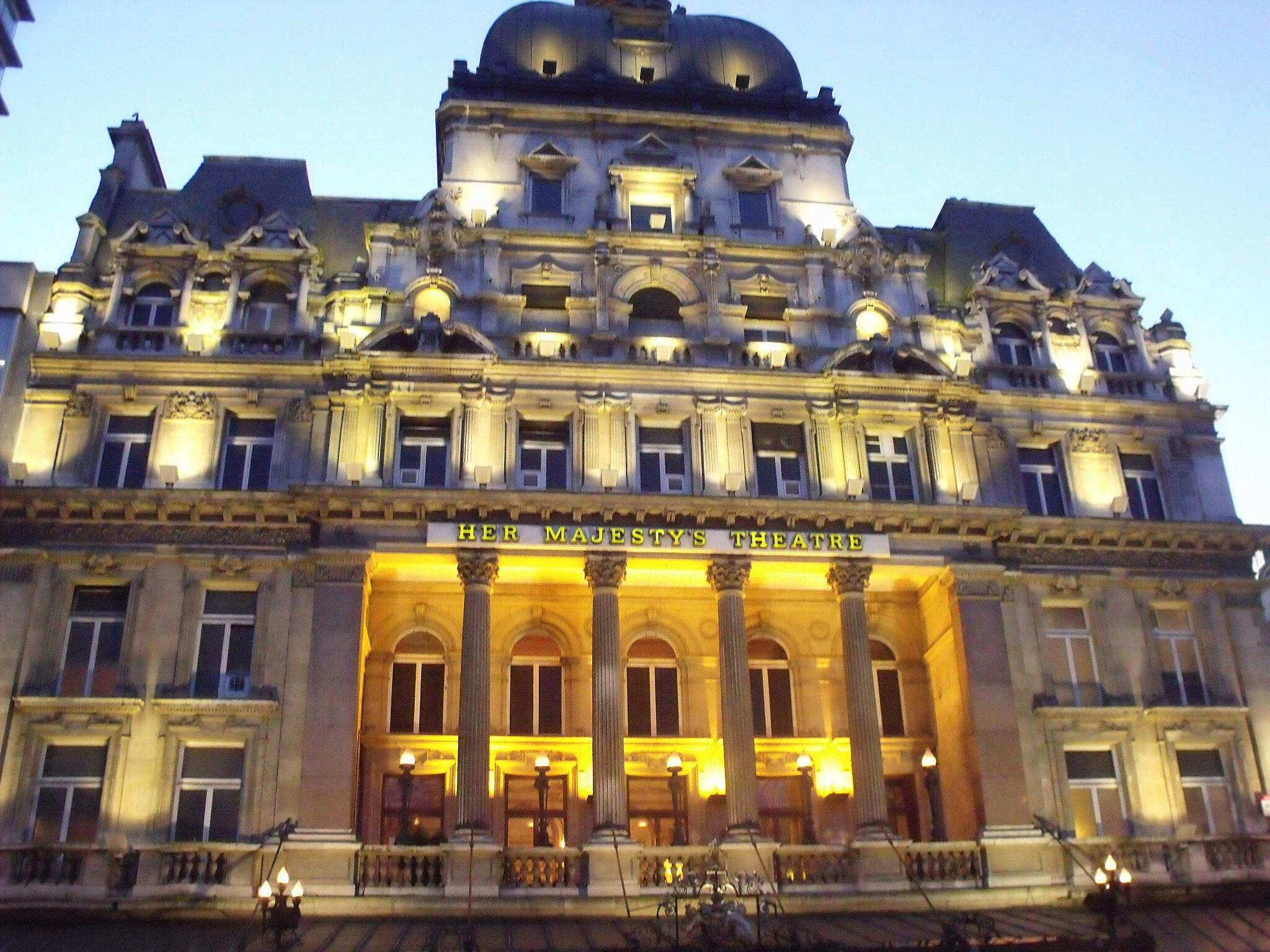 Facciata illuminata dell'Her Majesty's Theatre al tramonto, che mostra i dettagli architettonici e le colonne.