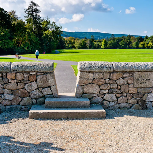 En person går bort på en stig genom en gräsbevuxen park, inramad av en stenmur märkt "The Wicklow Way".