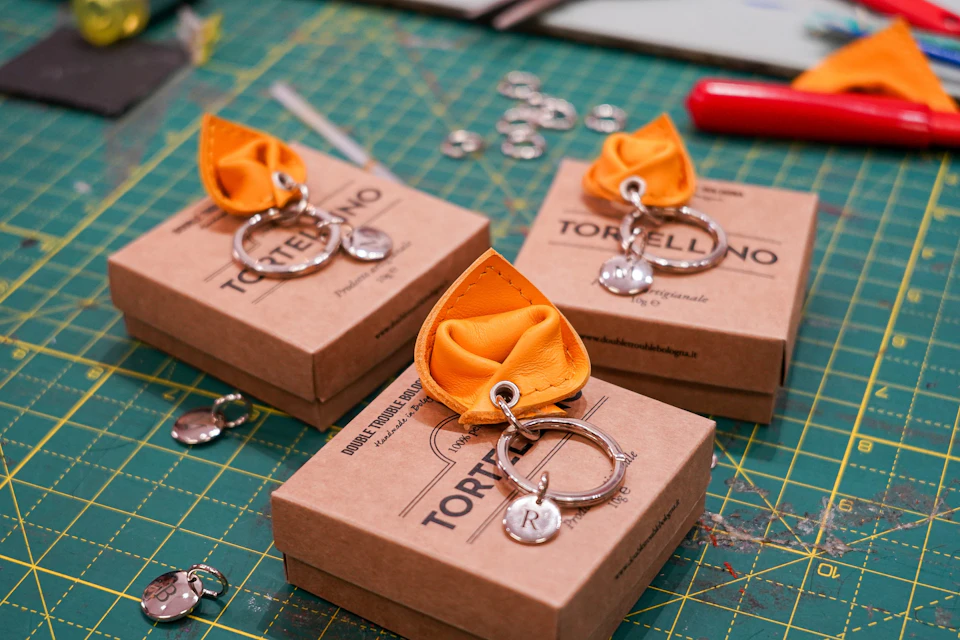 Three boxed tortellini-shaped keychains with yellow fabric and metal rings labeled "Tortellino" on a cutting mat.