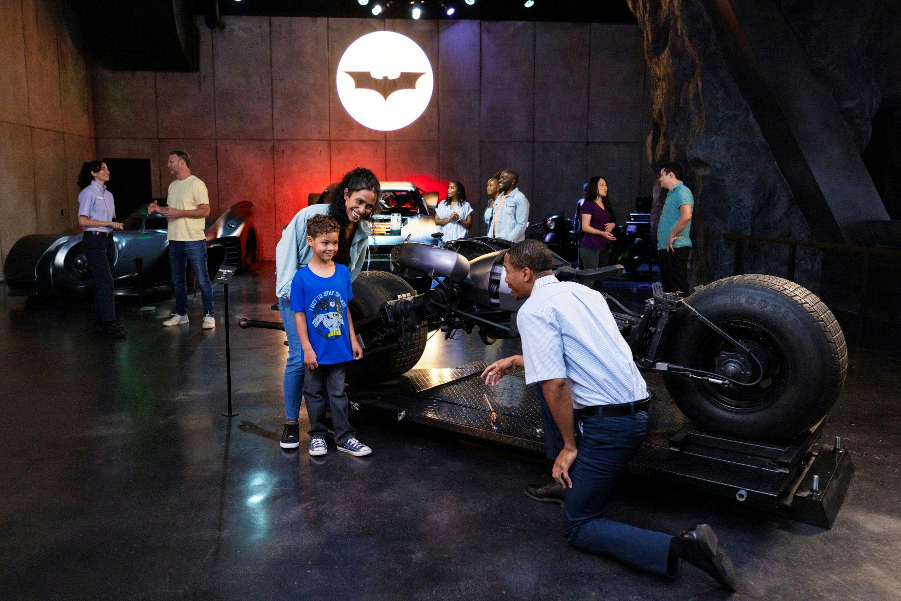 バットマンの展示室では、車や人々が交流し、薄暗い部屋の壁にバットのシンボルが投影されています。