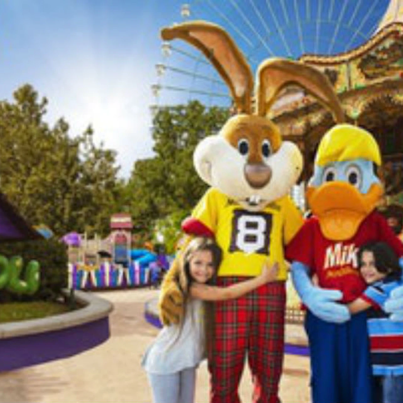 Two children smile and hug two costumed characters, a rabbit and a duck, in an amusement park near a carousel.