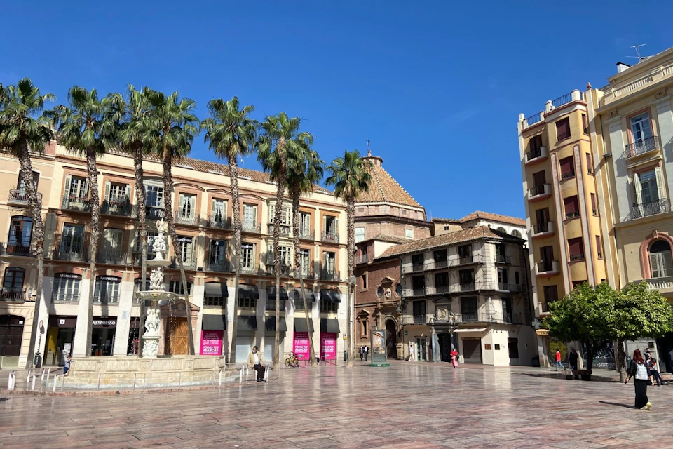 Sunny town square with palm trees, historic buildings, a fountain, and few pedestrians. Blue sky in the background.