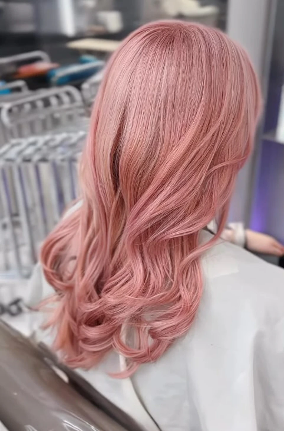 Wavy pink hair styled neatly, with a blurred background of metallic objects and a person in a light-colored outfit.