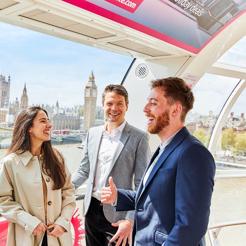 Tres personas sonriendo y hablando en el interior de una cápsula del London Eye, con el Big Ben y las Casas del Parlamento visibles al fondo.