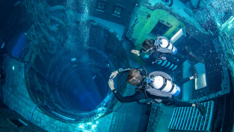 Two divers in scuba gear explore an underwater tunnel surrounded by submerged buildings and structures.