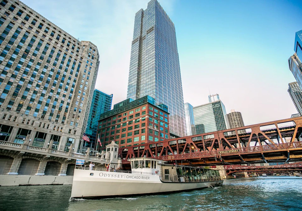 A boat named "Odyssey" travels on the Chicago River with tall buildings and a bridge in the background.