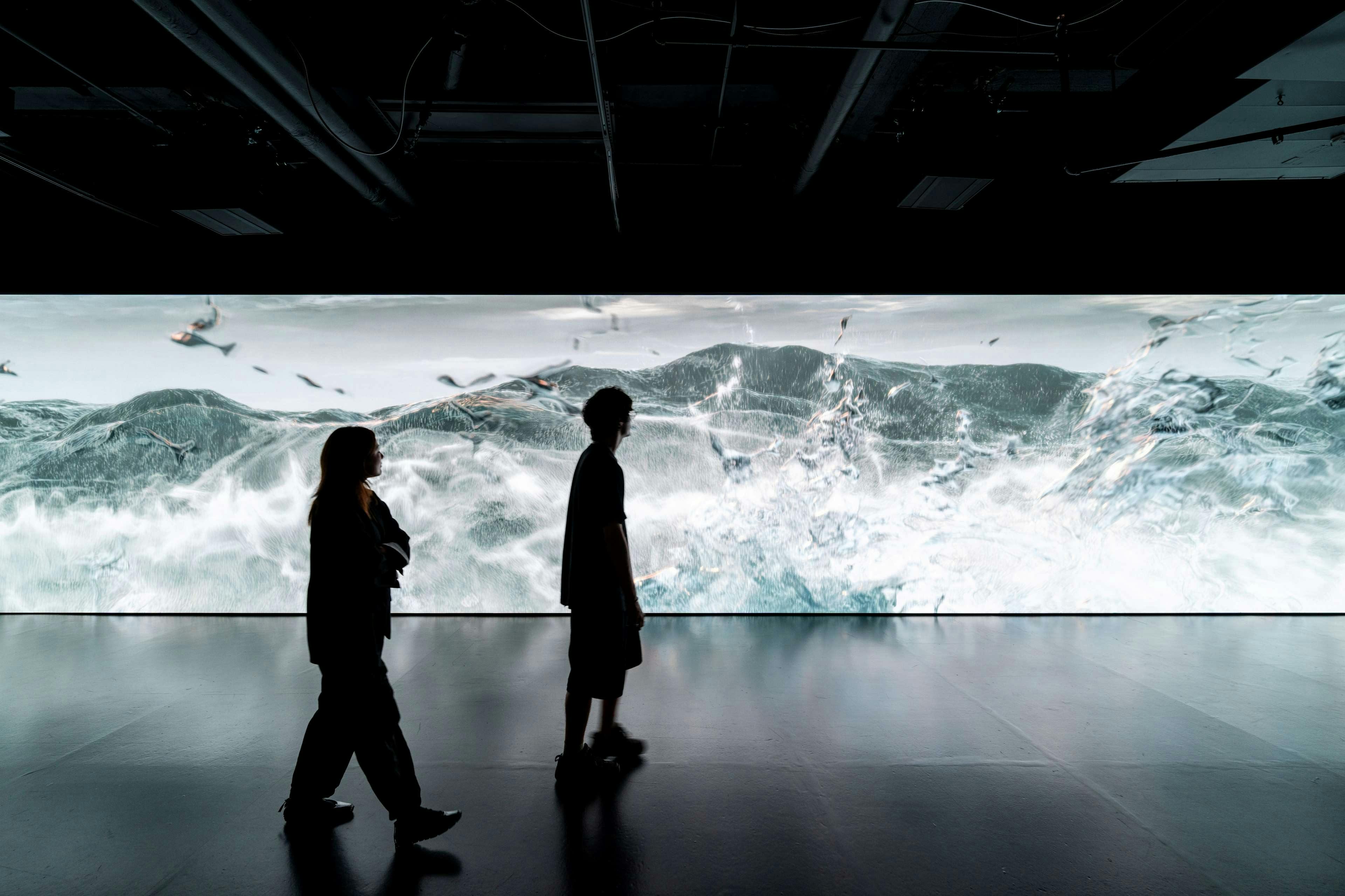 Zwei Personen gehen vor einem großen, beleuchteten Bildschirm, der eine Meereswelle in einem dunklen Innenraum zeigt.