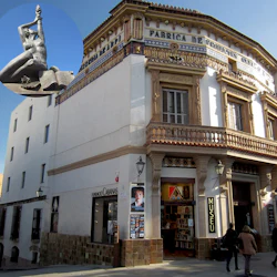 Corner building with ornate facade, bookstore entrance labeled "MUSEU," two people walking. Inset: close-up of a statue.