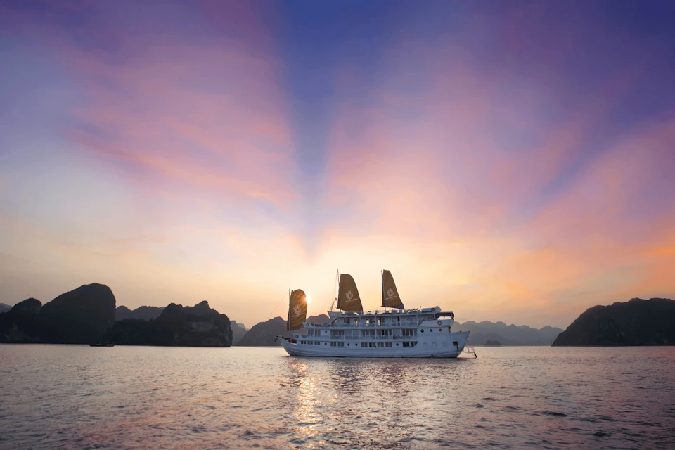 A large white ship with dark sails is on calm waters at sunset, with mountains silhouetted in the background.