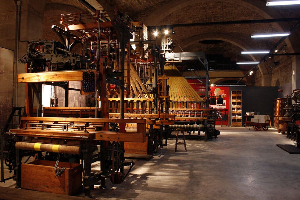 대형 산업용 직기 및 기계, 목재 기둥, 조명 기구, 아치형 벽돌 천장이 있는 객실입니다.