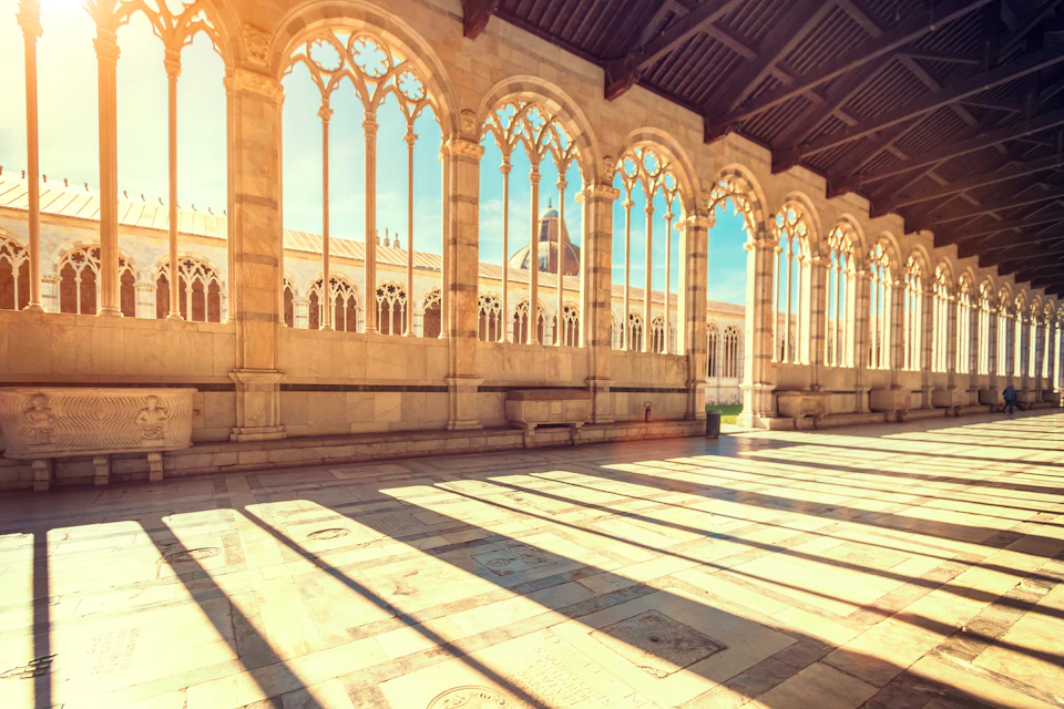 Sunlit medieval corridor with arched windows casting shadows on the stone floor. Roof beams and distant dome visible.