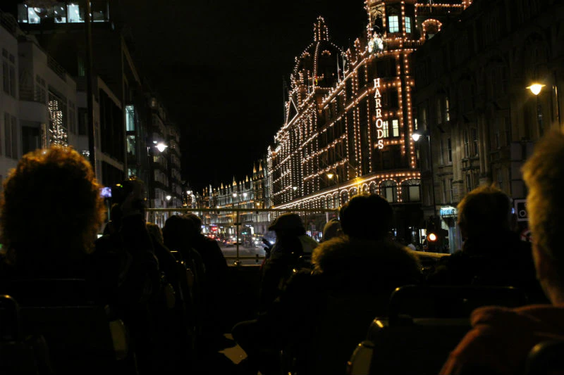 People on a nighttime city bus tour with view of an illuminated Harrods building in the background.