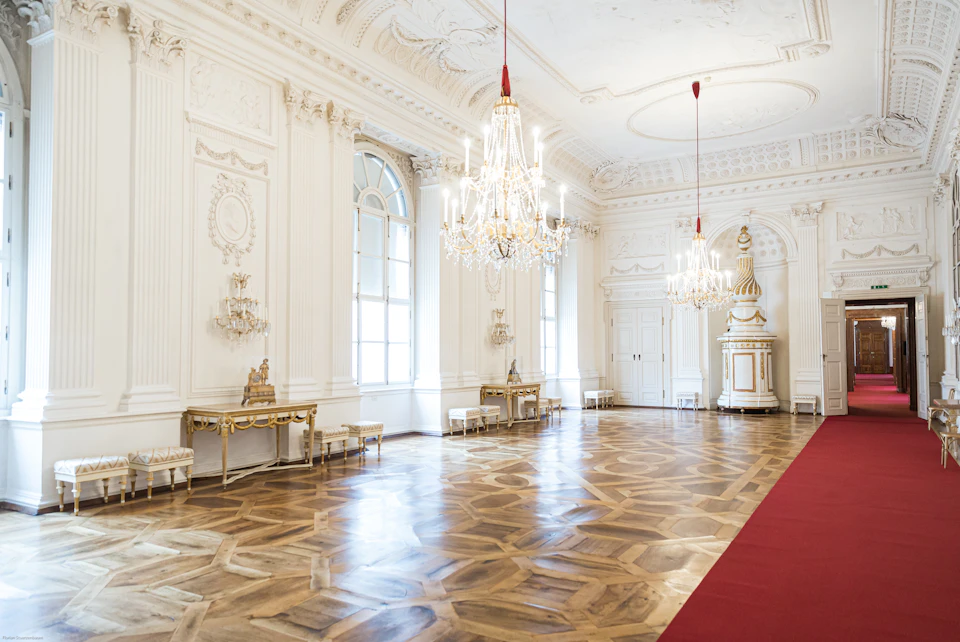 A spacious, ornate room featuring chandeliers, parquet flooring, white walls, furniture with gold detailing, and an aisle lined with red carpet.