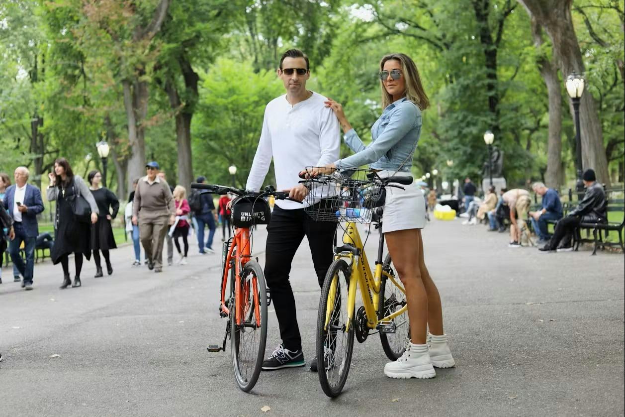A man and a woman stand next to their bicycles in a park, with people walking and trees in the background.