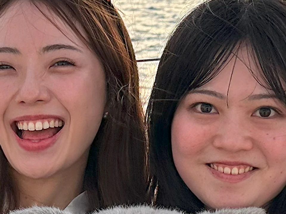 Two women smiling and posing with a koala plush toy on a boat, with a city skyline and a bridge in the background at sunset.