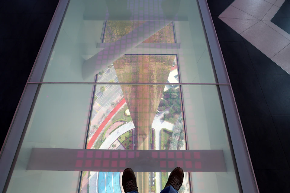 View through a glass floor showing a high-rise tower and landscaping below, with a person's shoes visible at the bottom.