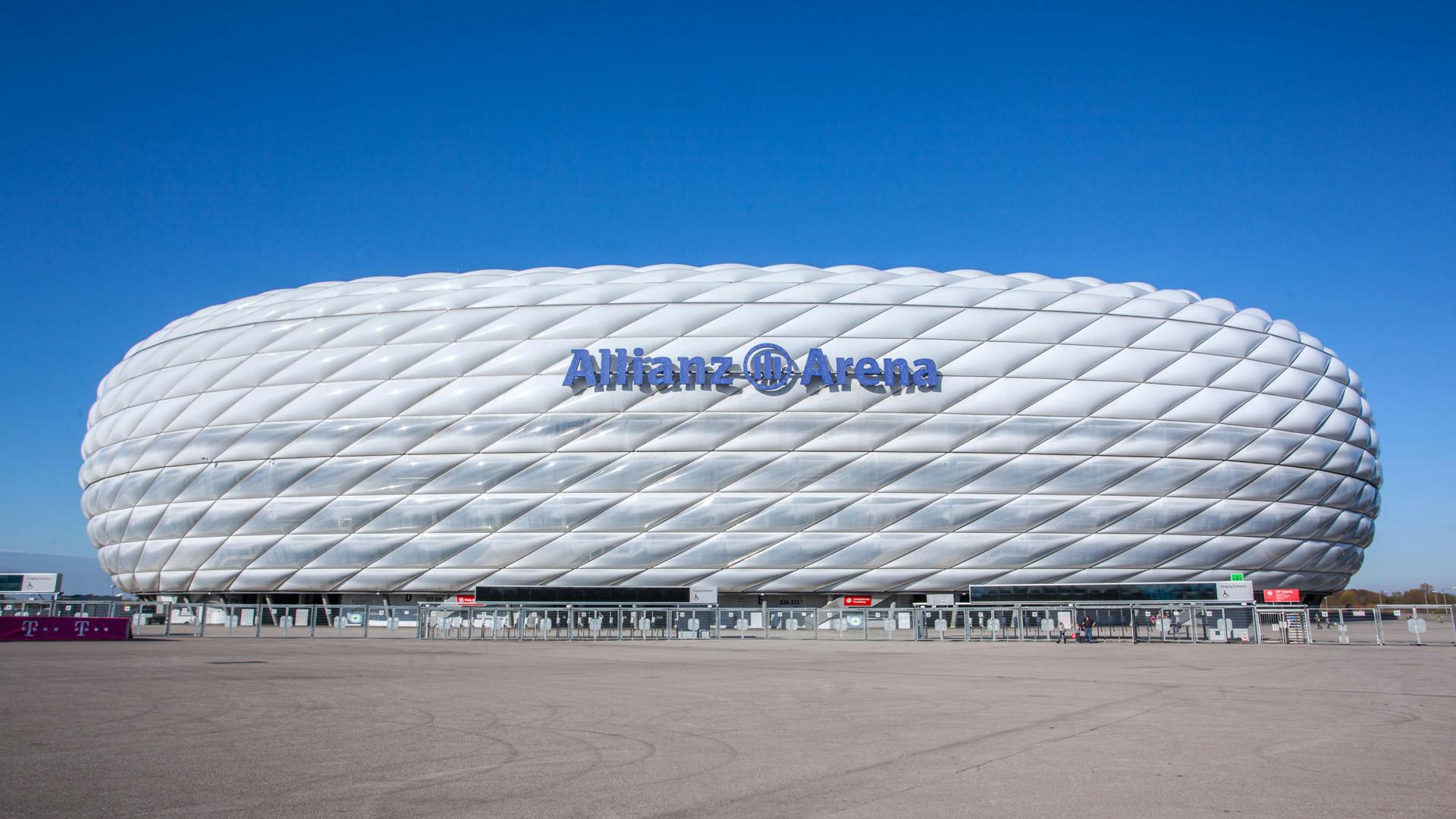 Velký bílý stadion označený „Allianz Arena“ proti jasně modré obloze s rozsáhlým prázdným chodníkem v popředí.